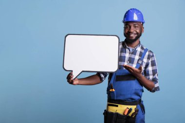 Smiling construction worker pointing to blank dialogue cloud with copy space, advertising mockup. Man holding speech bubble with message frame looking at camera, studio shot against blue background.