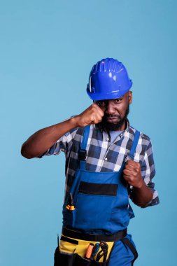 Aggressive handyman wearing coveralls trying to fight on camera, showing boxing fists with angry expression. African american builder with protective helmet in studio shot against blue background.