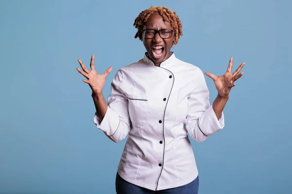 Angry african american female chef screaming loudly and waving her hands in a fit of rage in studio shot. Female cook in uniform looking frustrated at work against blue background.