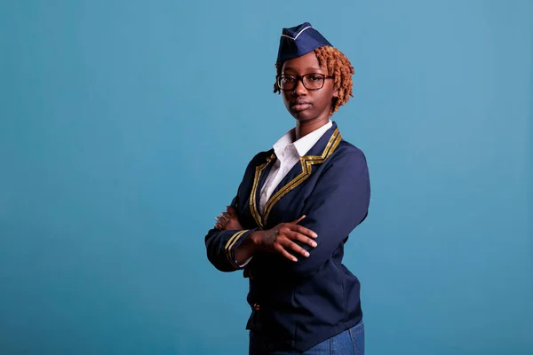 Close-up view of a flight attendant with arms crossed, tired from all the work in a studio shot. Stewardess in uniform and glasses appearing serious while looking at camera against blue background.