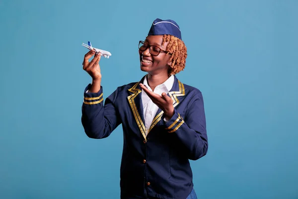 African american flight attendant in work uniform holding a model airplane. Stewardess showing commercial scale airplane with space for airline advertising in studio shot.