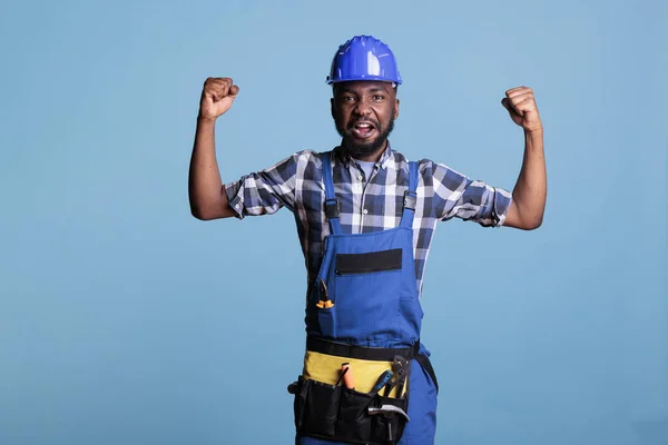 African american construction worker in coveralls showing his arm muscles in front of the camera. Construction worker in hard hat demonstrating his strength against blue background in studio shot.