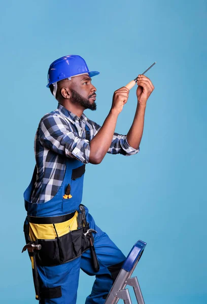 African American construction worker on ladder repairing something with screwdriver. Construction worker wearing uniform and hard hat against blue background in studio shot