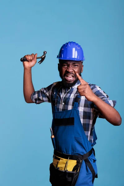 Angry construction worker angry and upset about overwork and long hours without rest. Frustrated upset person shouting aggressively while having an argument with employer against blue background.