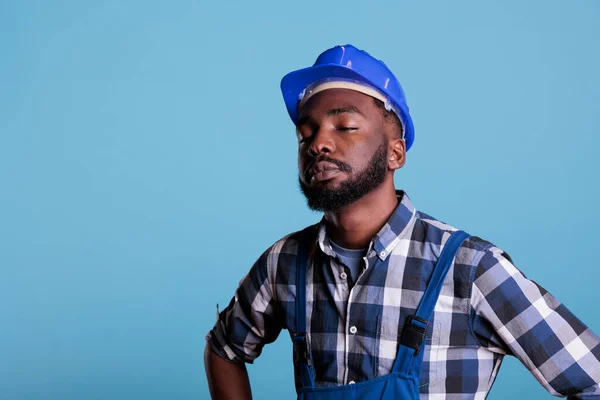 Tired exhausted construction worker in construction helmet and coveralls. African american employee with closed eyes feeling fatigued after hard work isolated on blue background.