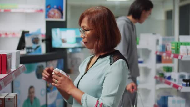 アジアの女性は棚の上に心臓病薬箱を読んで 医療薬を探しています 薬局で医薬品パッケージをチェックする女性の顧客 医療用品やビタミン — ストック動画