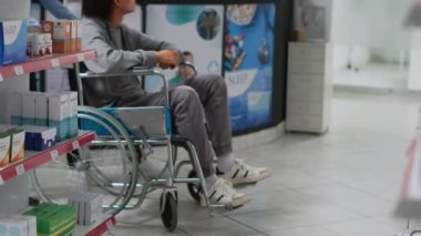Young man in wheelchair coming to pharmacy to buy drugs, receiving help from asian caretaker. People talking to consultant about medicaments and healthcare treatment in drugstore.