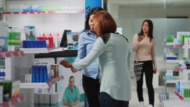 Asian women talking about medicaments in pharmacy, specialist giving box of vitamins to female client. People looking at drugstore shelves to find medicine and pills, pharmaceutical products.