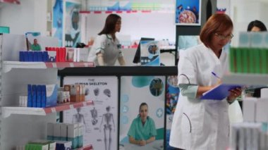 Asian pharmacist checking medicaments inventory in drugstore, looking at pills stock to give to customers. Pharmaceutical worker analyzing supplements and medical products, taking notes.