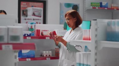 Female pharmacist arranging medical products on store shelves, preparing to help customers. Asian consultant looking at boxes of medicaments and vitamins, checking pills packages.