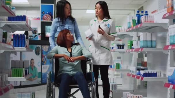 介護者と車椅子の女性ユーザーは薬や薬について薬剤師に尋ねます 薬局で処方薬を探しています ドラッグストアで障害者を支援するソーシャルワーカー — ストック動画