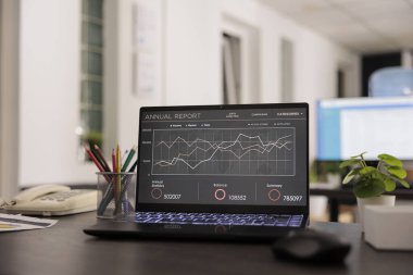 Inside of a startup companys modern office in the financial district, no people working in it. Wokplace with annual data report on portable computer screen on desk, employees on lunch break.
