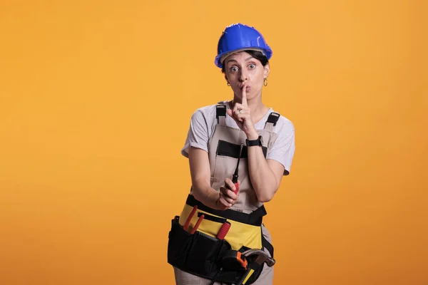 Female builder advertising hush secrecy sign, standing over yellow background and asking for privacy. Woman construction worker wearing uniform and helmet, doing mute confidential symbol.