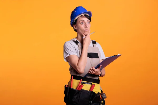 Female contractor analyzing building measurements on papers, looking at notes on clipboard. Construction worker measuring renovating project on yellow background, studio shot.