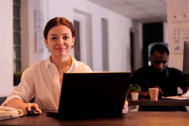 Smiling professional using laptop in office, looking at camera, woman typing on computer portrait. Corporate worker browsing website at workplace desk, analyzing marketing report