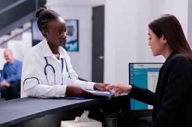 Receptionist helping physician doctor with checkup visit appointments while working at hospital registration desk in waiting area. Medic reviews the patients medical history before their consultation.