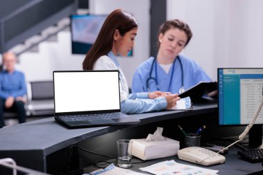 Laptop computer with isolated white screen standing on registration counter in hospital waiting area. Medical team working with sick patients during checkup visit, offering health care treatment
