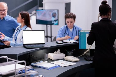 Receptionist talking with nurse discussing medical report during checkup visit consultation in hospital waiting area. Laptop computer with white isolated screen standing on registration counter desk