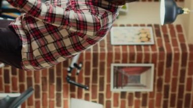 Vertical video: Modern male person doing spring cleaning session at home, using vacuum cleaner to clean up apartment. Young cheerful man vacuuming wooden living room floors, using washing solution.