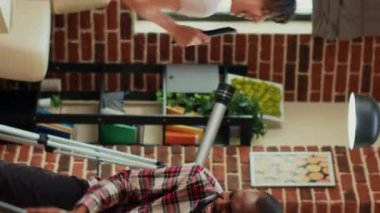 Vertical video: Interracial partners having fun cleaning room with vacuum cleaner and mop, dancing and listening to music. Modern cheerful people enjoying spring cleaning, vacuuming apartment floors.