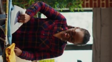 Vertical video: African american woman having fun ironing clothes in living room, listening to music and dancing while she does housework. Modern girl showing dance moves and being silly, enjoyment.