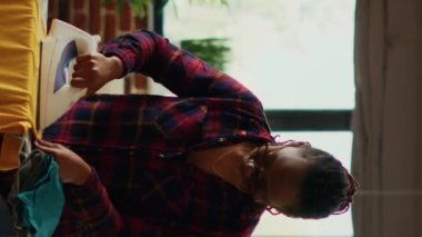 Vertical video: African american woman using steam iron and ironing board, smoothing out creases on clothes. Female person doing household chores for spring cleaning, using laundered clothing.