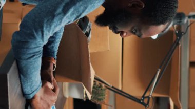 Vertical video: African american man analyzing stock and orders, doing inventory on laptop at warehouse space. Male employee using logistics and financial planning to send merchandise for business