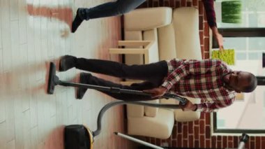 Vertical video: Young happy couple listening to music and washing floors in apartment, doing silly dance moves. Cheerful people doing spring cleaning and dancing, mopping tiles. Handheld shot.