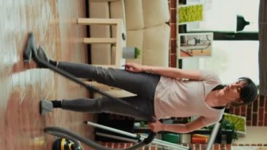 Vertical video: Young woman sweeping dirt with mop in living room, mopping wooden floors and cleaning apartment. Casual female person washing tiles with cleaner, being focused on housework or chores.