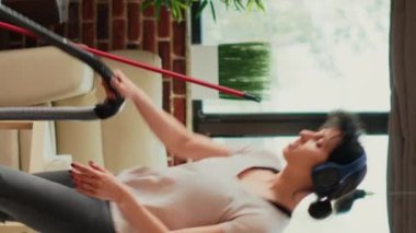 Vertical video: Female person listening to music on headset and vacuuming floors, having fun with vacuum cleaner. Young modern woman feeling cheerful while she does spring cleaning household.
