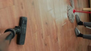 Vertical video: POV of person using vacuum cleaner to tidy up apartment, cleaning dirt or dust in household. Young adult vacuuming wooden floors, doing chores with life partner cleaning furniture