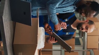 Vertical video: Diverse startup partners doing quality control before shipping goods order, packing merchandise in cardboard boxes. Workers team preparing delivery and shipment in warehouse storage