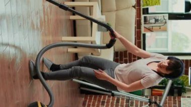 Vertical video: Female adult listening to music on headset and vacuuming floors at home, doing spring cleaning work in apartment. Young wife singing and doing dance moves while she uses vacuum.