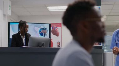 Male doctor asking receptionist about medical appointments, sitting at lobby counter. General practitioner and employee analyzing checkup reports befoe consulting people. Handheld shot.