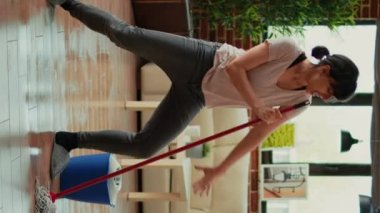 Vertical video: Happy funny person mopping floors with all purpose cleaner and dancing, spring cleaning. Young cheerful housewife using mop to sweep dirt and clean mess, listening to music at home.