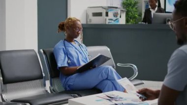 Female nurse looking at medical reports in waiting area, sitting in facility lobby. Young healthcare assistant looking at checkup forms before having consultation appointment in health center.