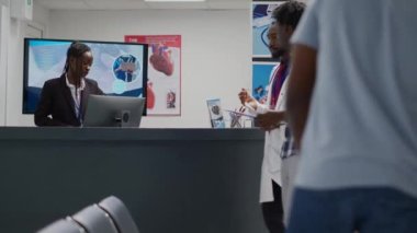 Professional medic giving advice to sick patient in facility hallway, man approaching reception counter to ask receptionist about insurance support. Young adult finishing consultation visit.