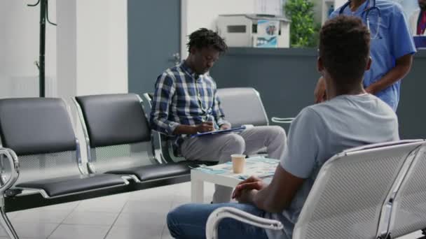 アフリカ系アメリカ人患者の健康診断報告書に記入し 病院の待合室のロビーに座っている 一般開業医との約束に出席する前に 医療用紙ファイルを読む男性 — ストック動画