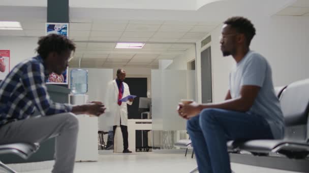 アフリカ系アメリカ人 病院のロビーで待っている病気の患者との検査を行う医療スタッフ 予約に出席する前に 待機エリアに座っている男性のグループ 三脚ショット — ストック動画