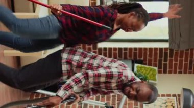 Vertical video: People of african ethnicity being funny and cleaning in living room, using mop to wash dirt and vacuum to clean floors. Young partners dancing and doing spring cleaning. Handheld shot.
