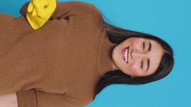 Vertical video: Professional housekepper pointing and showing cleaning advertisement or isolated text, standing on blue background in studio. Housewife worked hard to maintain a clean home, using