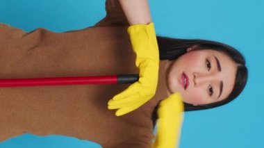 Vertical video: Tired exhausted maid falling asleep on broom while cleaning house, being overworked in studio over blue background. Sleepy professional housekepper was responsible for the houseclean