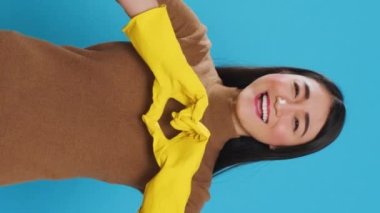 Vertical video: Happy asian maid wearing yellow gloves while doing heart shape in front of camera after finishing cleaning houses for customers. Housekeeper primary responsibilities is to maintain