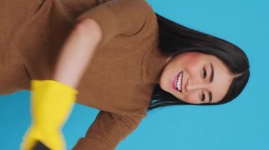 Vertical video: Happy asian maid wearing protective gloves while cleaning house using sponge and detergent spray, standing in studio over blue background. Housekeeper is an expert in cleaning