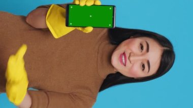 Vertical video: Asian homemaker holding chroma key mock up mobile phone with greenscreen display while talking with remote client during videocall meeting. Cheerful maid was skilled in all aspects of