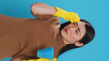 Vertical video: Pensive thoughtful maid holding cup of coffee standing over blue background in studio, thinking at solution to solve cleaning problem. Housekepper providing top-notch cleaning services
