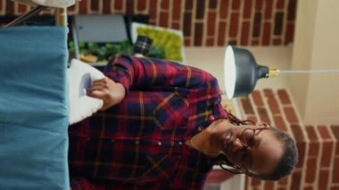 Vertical video: Modern wife getting tired and angry about doing chores alone, ironing clothes with steam tool in apartment. Young person being unhappy and irritated about housework. Handheld shot.