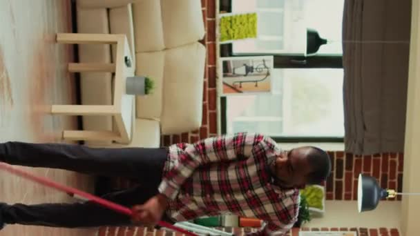 垂直视频 年轻夫妇用吸尘器和家具抛光剂清洁客厅 女人用各种目的的清洁剂清扫架子上的灰尘 非洲裔美国人在家里吸尘木地板 — 图库视频影像