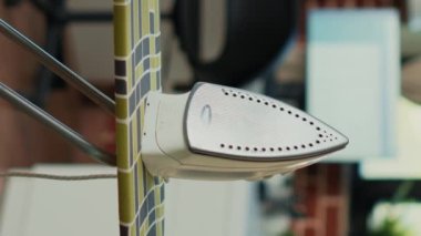 Vertical video: Empty household with iron and ironing table, big pile of washed clothes on board to smooth out creases and wrinkles. Living room with home appliances and tools to iron clothing, house