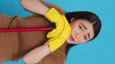Vertical video: Tired housekepper falling asleep on broom while cleaning client home, being overworked in studio over blue background. Sleepy maid wearing rubber gloves while being was responsible for